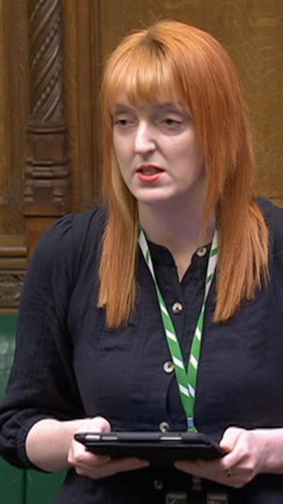 A woman stood up in Parliament, holding an iPad and wearing a blue shirt and green lanyard. She looks serious, and has ginger hair and pink lipstick.