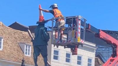 Man standing on a cherry picker removes cone from the top of a statue of Oliver Cromwell.