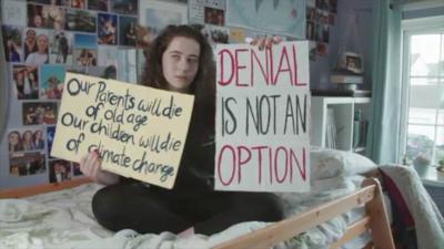 A girl sits on a bed holding signs saying "Our Parents will die of old age, our children will die of climate change" and "Denial is not an option".