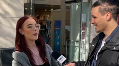 Journalist Tyler Edwards smiles and holds a BBC microphone towards a women with red hair and glasses
