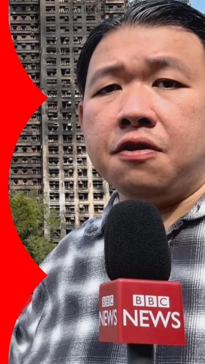 A man stands in front of a burnt out building holding a bbc branded mic