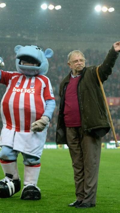 A football mascot wearing a red and white striped shirt. The mascot is a blue elephant. He is next to a man in kahki trousers, green jacket and red jumper and glasses with grey hair