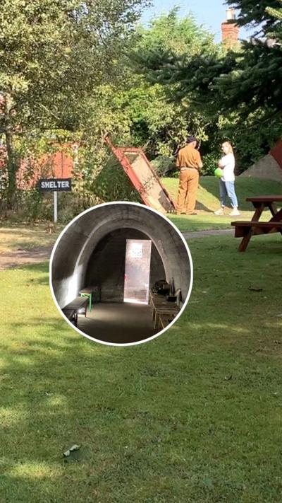 A man in brown army uniform is pictured talking to a woman who is wearing jeans and t-shirt. They are standing next to a sign saying shelter in white text on a black background. The entrance to the shelter is behind them and looks like a doorway at a 45 degree angle to the grassed lawn it leads down into. 