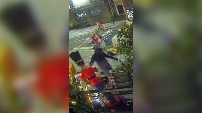 A person wearing dark coloured clothing, riding a bike is grabbing a large nutcracker statue with their right hand. The statue is facing away from the camera and it has a red jacket and black hat. There is a road behind the dark clothed person and a Christmas tree is on the right.