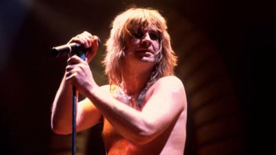 Ozzy on stage in 1982, with no top on, in front of a mic.