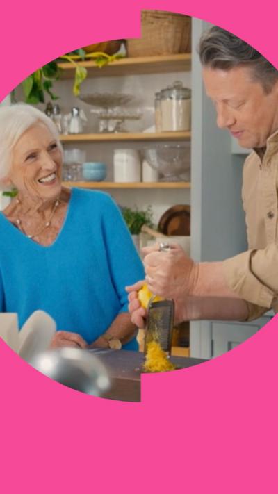 Mary Berry smiles at Jamie Oliver in a kitchen. He is grating a lemon on a chopping board