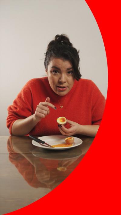 A woman with brown hair in a messy bun sits at a table with a knife, plate and a soft-boiled egg. She is wearing a an orange jumper and pointing to the inside of half the egg, which she is holding.