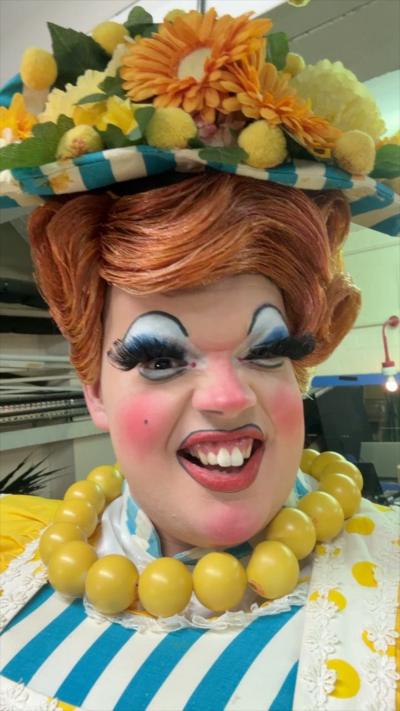 A pantomime dame wearing a big yellow necklace smiles at the camera. 