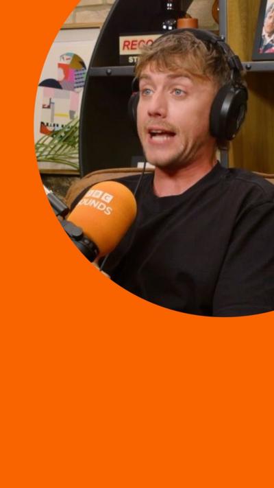 A man, Roman Kemp, wears a black top and sits on a sofa in front of a Sounds branded microphone