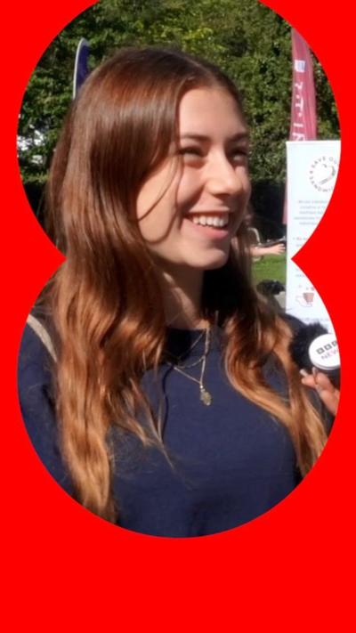 A young woman with long brown hair and a blue jumper smiles at someone off camera