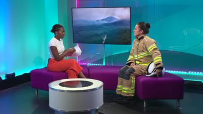 Two women sitting on a purple sofa speaking. One is wearing a firefighter uniform.