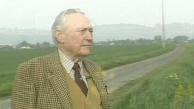 Richard Todd stand in a tweed jacket, mustard wastecoat maroon tie and white checked shirt, look contemplatively out over a field in Normandy.