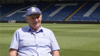 Frank Rothwell is wearing a light blue shirt and a navy flat cap. He is sat next to the Oldham Athletic pitch. 