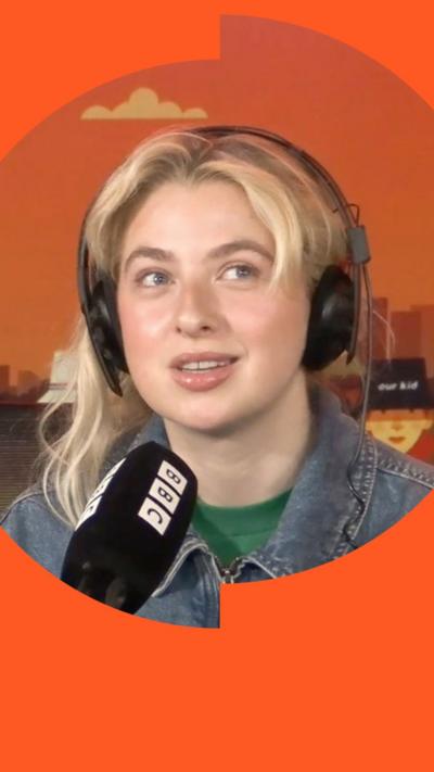 Anaïs Gallagher has long blonde hair and she is wearing headphones and sitting in front of a BBC-branded mic