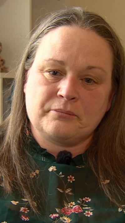 A woman with long light brown hair. She is wearing a dark green top with flowers on it 