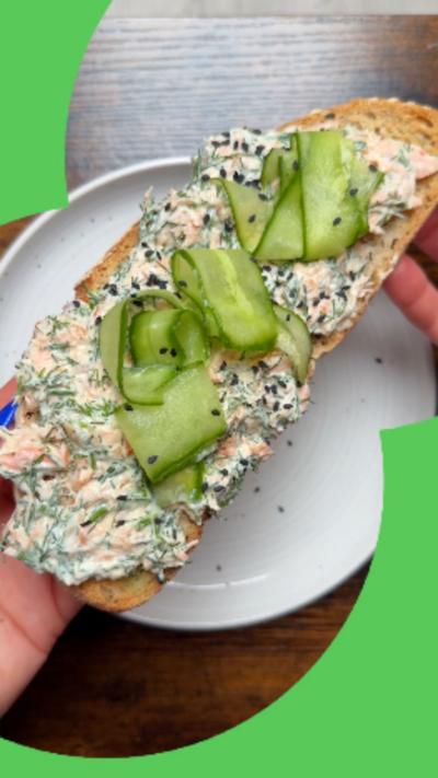 Smashed salmon on toast with ribbons of cucumber on top, seen from above with a person's hands lifting it from a plate.