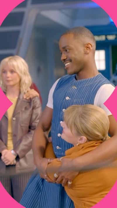 Ncuti Gatwa smiles while being hugged by a co-star on the set of Doctor Who. He wears a striped denim top and skirt
