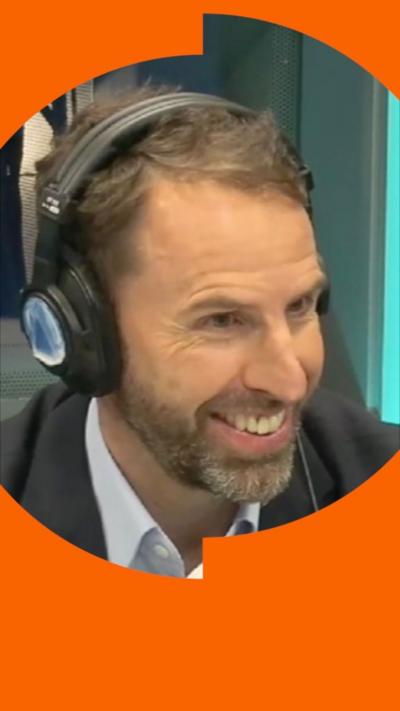 A man, Gareth Southgate, wears a shirt and suit jacket. He has headphones on and is smiling.