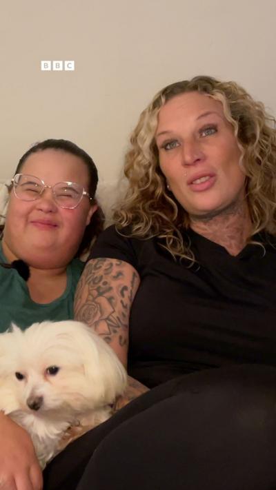 A woman with curly hair sits next to another woman wearing glasses. The woman wearing glasses is smiling. There is a white dog in her lap.