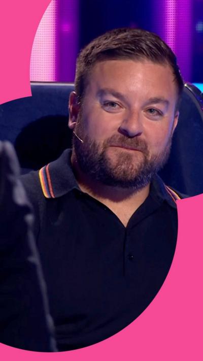 Alex Brooker looks towards the camera. He is wearing a polo shirt and sits in a chair. He has short hair and a short beard