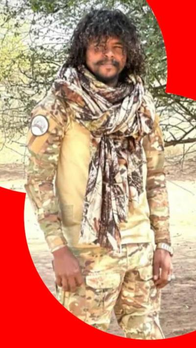 A man with dark curly hair wearing camouflage clothing standing outside next to a tree. 