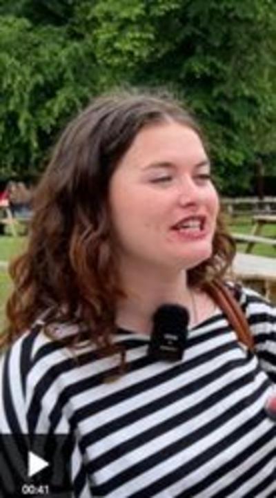 A young woman looks to the right of the camera, like she is talking. She has long dark brown hair, and is wearing a black and white striped t-shirt and a clip-on microphone. Behind her are green trees and grass, and some picnic benches.