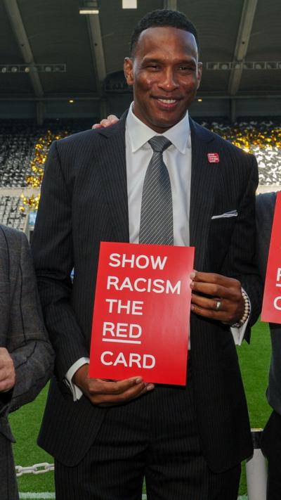 Shaka Hislop stands in front of St James' Park's East Stand in a dark pinstripe suit, holding a red sign that reads Show Racism the Red Card. 