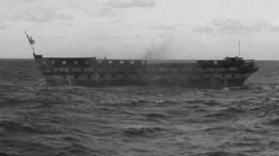 Black and white image of HMS Implacable out at sea, with bottom blown out and back broken, smoke rising from it, beginning to sink. A flag is visible at one end.