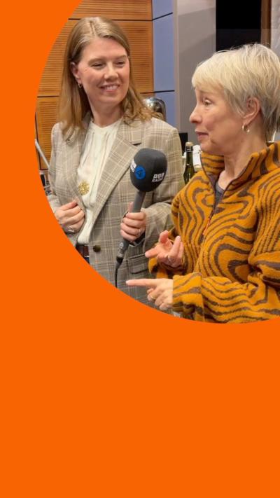 Two women in a radio studio. Felicity speaks into a mic while Nuala looks at her and smiles 