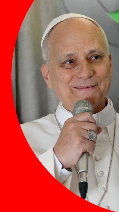 Pope Leo is dressed in white papal garments and holding a microphone.