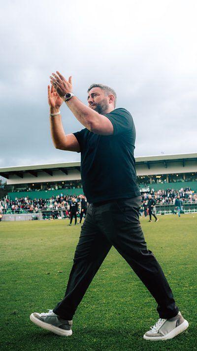 Lee Johnson on the pitch in Belgium, wearing green, with his hands in the air