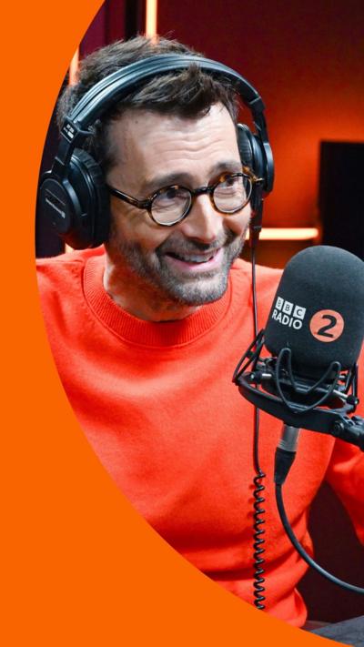 David Tennant smiles. He is wearing an orange jumper and wears black headphones. He sits in-front of a Radio 2 microphone.