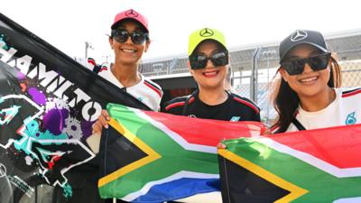 Three Formula 1 fans wear Mercedes branded caps and hold up South African flags and a banner reading 'Hamilton 44'