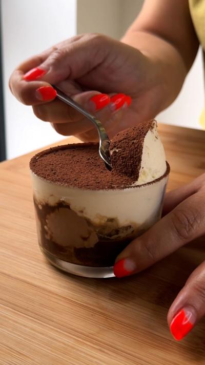 A pair of hands with neon coral-coloured fingernails scoop tiramisu out of a small dish