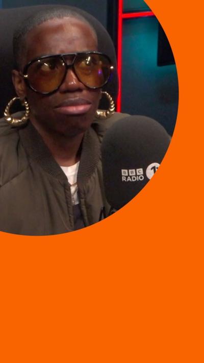 Candice sits behind a mic in a studio. She has short black hair and wears big black sunglasses and gold hoop earrings