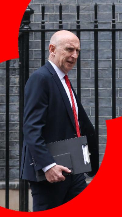 John Healey wears a dark suit with a white shift and red tie walking with a black folder under his arm.