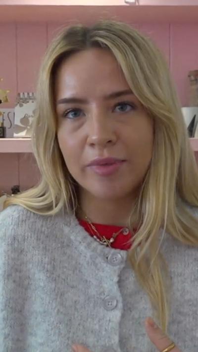 A blonde woman in a grey cardigan looking straight ahead in a shop with pink walls behind her
