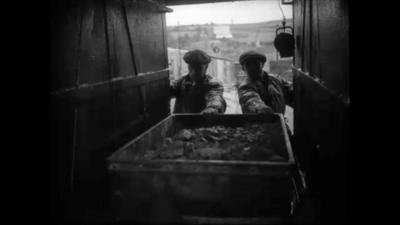 Two men pull a mine cart full of tin out from a mine shaft