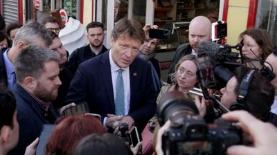 Reform UK deputy leader Richard Tice in a dark suit with light blue tie in among a crowd of press reporters in front of a shop.