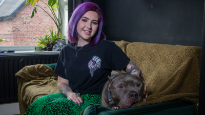 Purple-haired Lainya and her XL Bully, Smokey, sat on a green sofa with a light brown blanket draped over it.