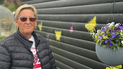 Hamilton resident Sandra Panton - a blonde woman in a black jacket, standing next to a black wooden shed with a hanging basket of purple flowers