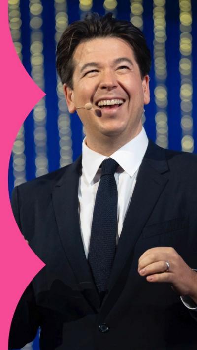 Michael McIntyre has black hair, is wearing a black suite with a purple shirt. He is smiling and holding cue cards