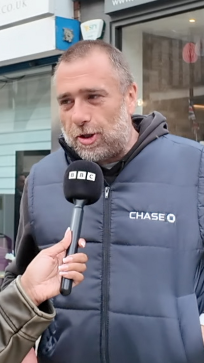 A man in a padded vest outside a hairdressing shop in a busy urban street.