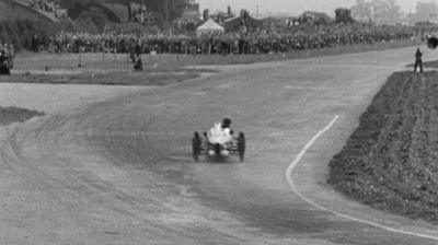 Black and white image of a racing car bearing down a wide open race track, having just taken a corner in the foreground.. Up ahead a steward is standing at the right hand side of the track in the distance