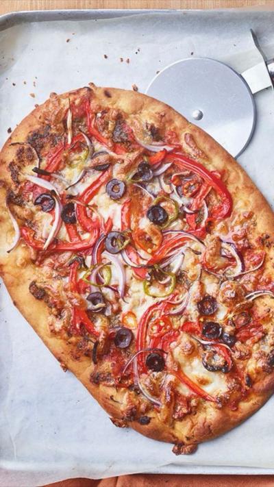 Top down view of a pizza in a baking tray with olives, red pepper and onions on. A pizza cutter is at the side.