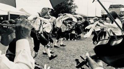 A black and white photo of Morris dancers in traditional dress and people playing the flute and violin.