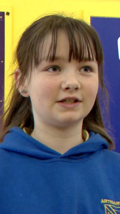 A child with long brown hair, a fringe and blue hoody with has her yellow crest on it 