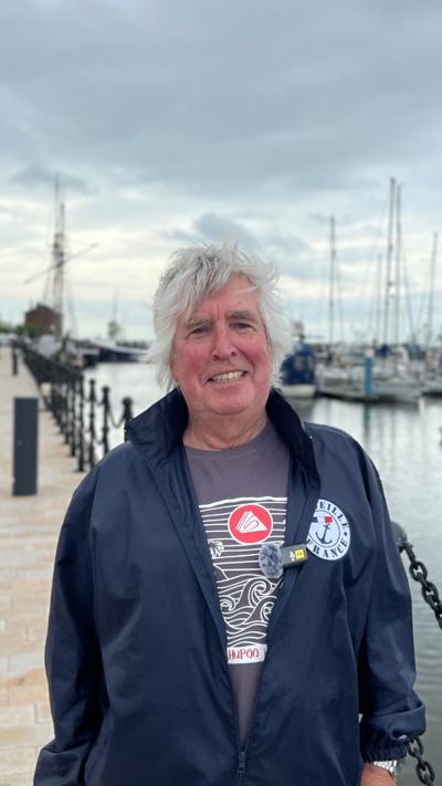 A man with white hair and a blue jacket smiles as he stands in front of a marina with yachts in the background.