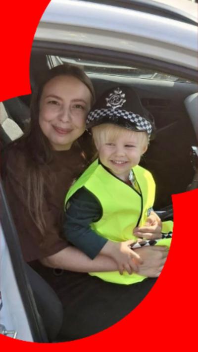 Small child wearing a police hat and high-vis vests sat on his mum's knee. They are sat on the driver's seat of a car with the door open. The woman has long dark hair.