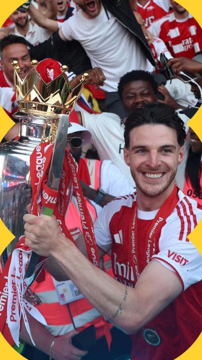 A man with short dark hair wearing a red and white football kit holds a trophy